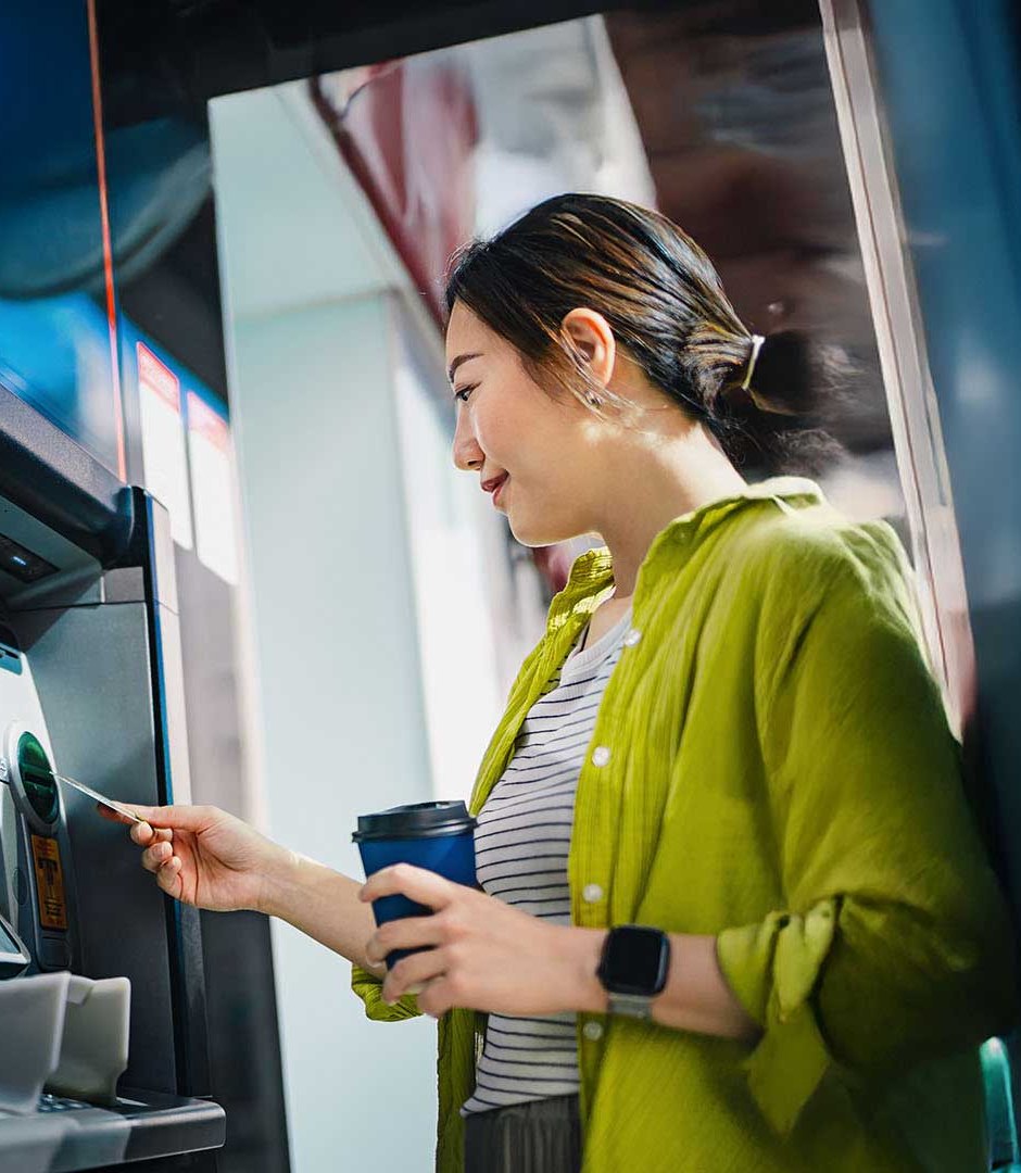 A woman holding a coffee cup inserts a card into an ATM